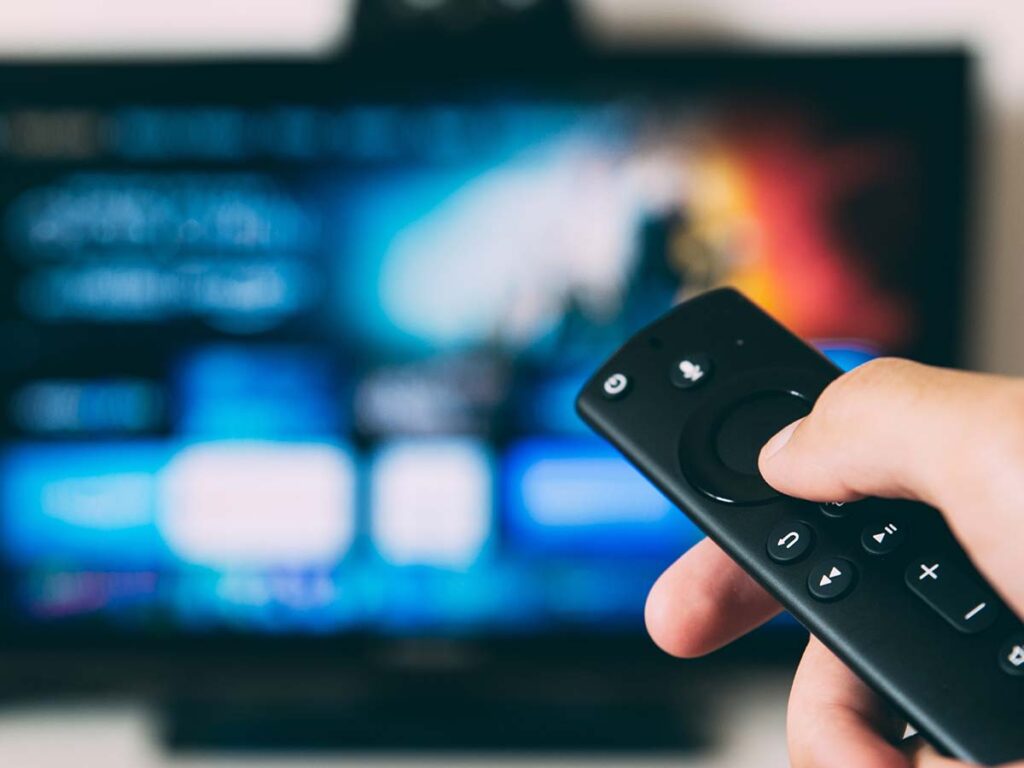 A closeup of a hand holding a remote control with a blurred television in the background