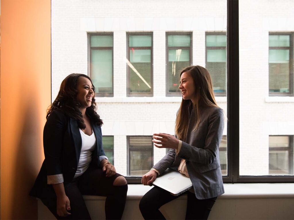 Two women sitting on a window sill talking.